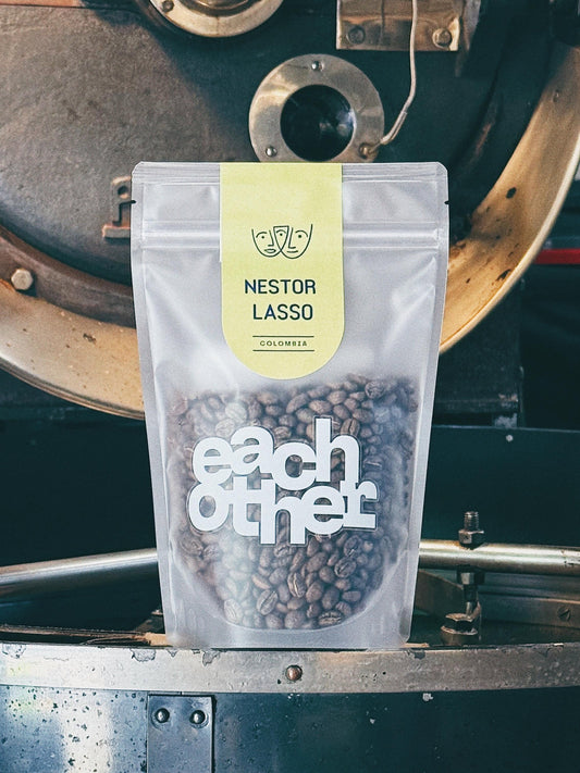 A stand-up pouch of Nestor Lasso Colombia coffee beans from Eachother, placed on the rim of a vintage coffee roaster. The pouch is clear, showing the dark roasted coffee beans inside, with a light yellow label at the top and bold white Eachother logo.