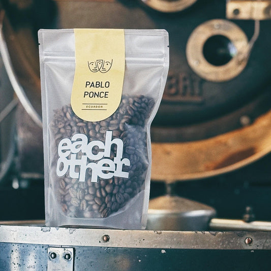 A stand-up pouch of Pablo Ponce coffee beans from Eachother, featuring a cream label and white logo, set against a blurred vintage coffee roaster background.