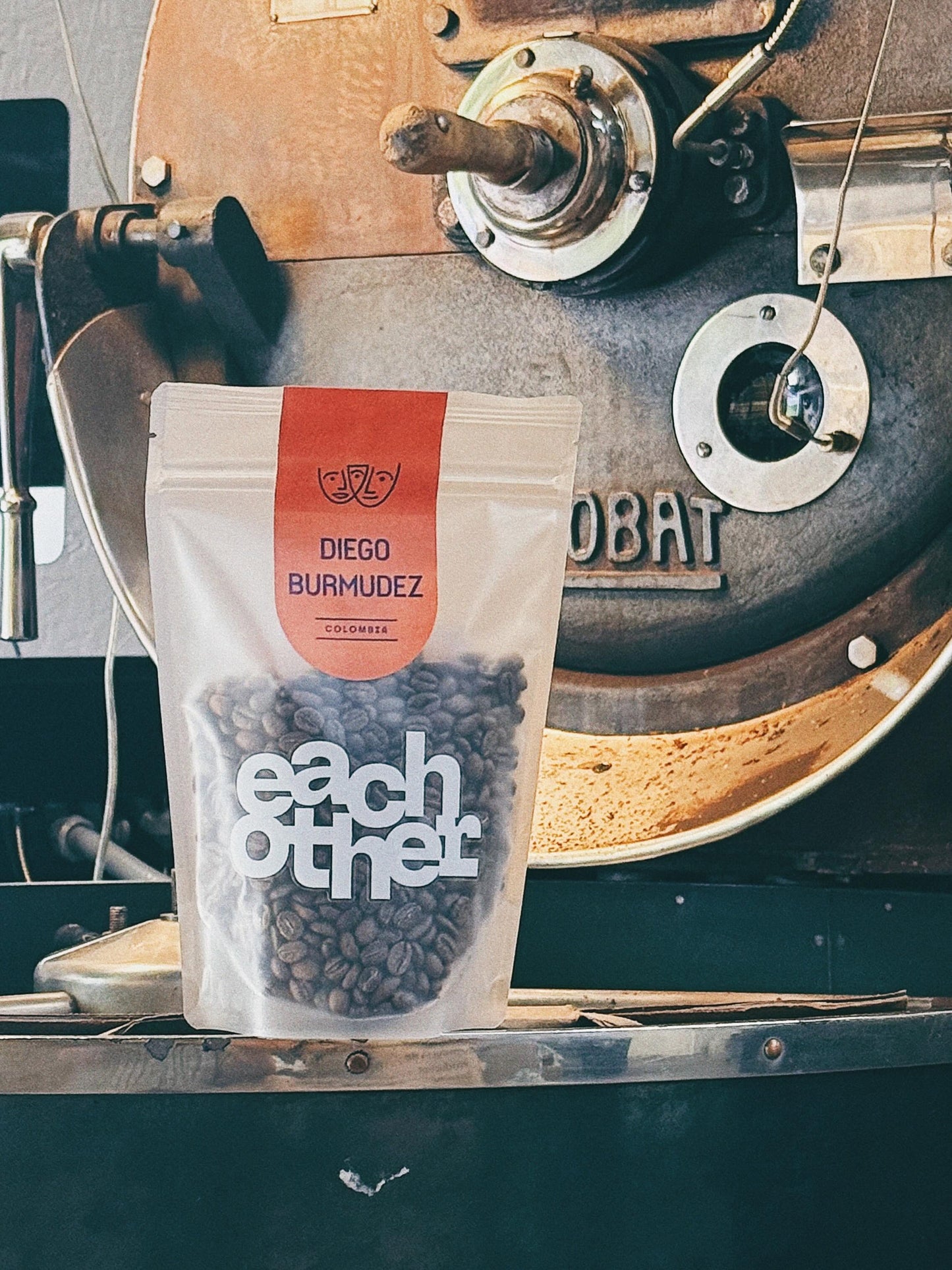 A bag of Diego Burmudez coffee beans by Eachother, placed in front of a vintage coffee roaster.