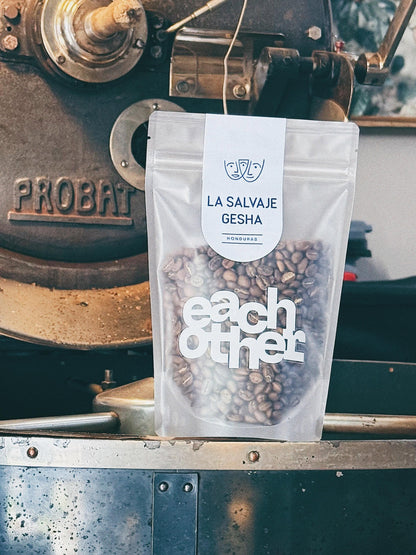 A stand-up pouch of coffee beans labeled La Salveja Gesha stands in front of a vintage coffee roaster. The pouch is clear enough to show roasted coffee beans inside, with a pale blue label at the top and a white Eachother logo in the middle.