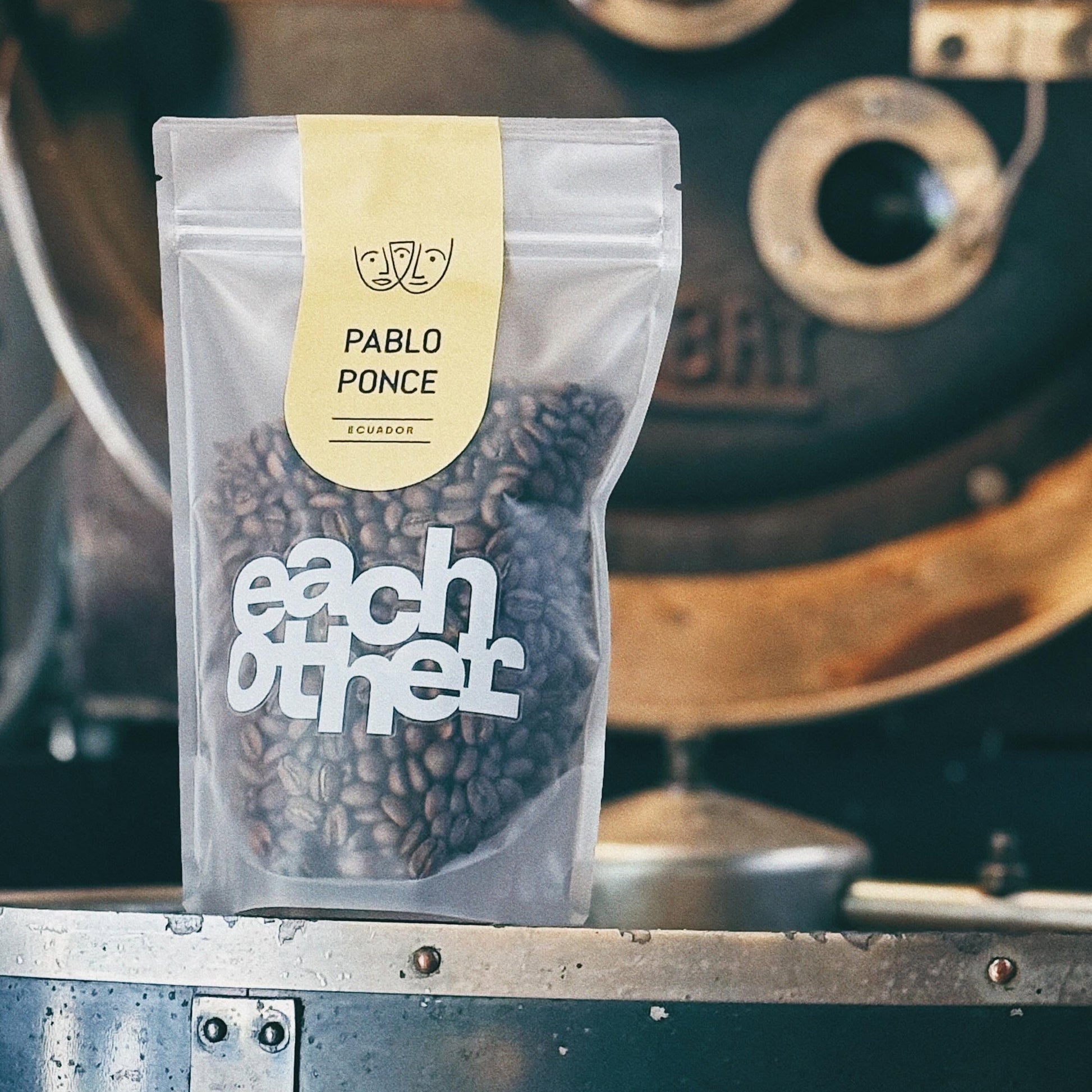 A stand-up pouch of Pablo Ponce coffee beans from Eachother, featuring a cream label and white logo, set against a blurred vintage coffee roaster background.